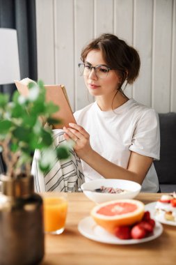 Evde rahat bir odada kahvaltı ederken kitap okuyan güzel odaklanmış bir kadının fotoğrafı.