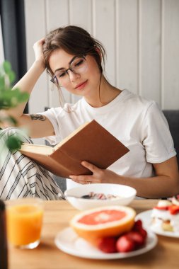 Evde rahat bir odada kahvaltı ederken kitap okuyan güzel odaklanmış bir kadının fotoğrafı.