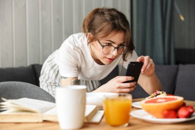 Güzel bir kadının evde rahat bir odada kahvaltı ederken cep telefonuyla fotoğraf çekmesi.