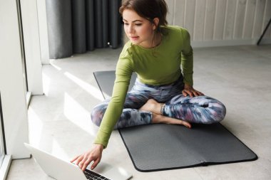 Odaklanmış esmer sporcunun, rahat bir odada minder üzerinde çalışırken dizüstü bilgisayar kullanmasının fotoğrafı.