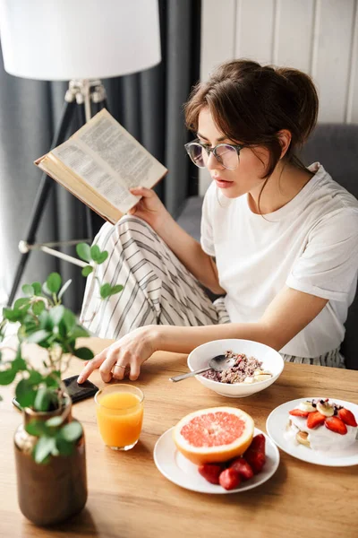 Photo Young Woman Reading Book Using Cellphone While Having Breakfast — Stock Photo, Image