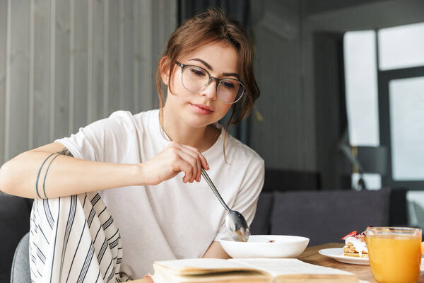 Photo of young thinking woman reading book while having breakfast in cozy room at home