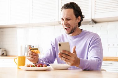 Çekici mutlu genç adam mutfak masasında oturuyor, kahvaltı yapıyor, mısır gevreği yiyor, cep telefonu ve kredi kartıyla online alışveriş yapıyor.
