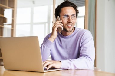 Çekici, neşeli genç adam mutfak masasında oturuyor, dizüstü bilgisayarla çalışıyor, cep telefonuyla konuşuyor.