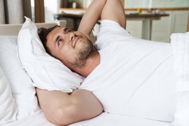 Yatakta yastığa uzanmış, ellerini uzatarak gülümseyen genç adam.