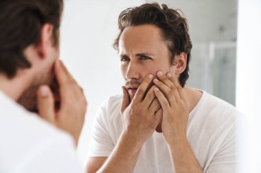 Çekici endişeli genç adam banyo aynasının önünde durmuş, yüzünü inceliyor.