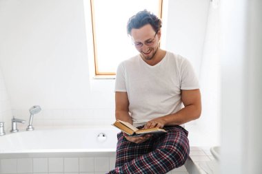 Pijama giymiş, küvette oturmuş kitap okuyan gülümseyen genç adam.