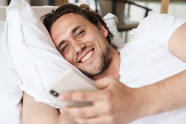 Yatakta yastığa uzanmış gülümseyen genç adam, cep telefonu kullanıyor.
