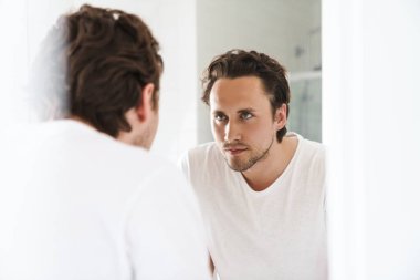 Çekici, kendine güveni olan genç bir adam banyo aynasının önünde duruyor ve kendine bakıyor.