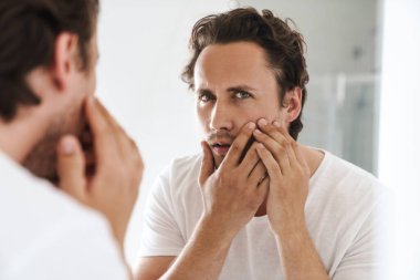 Çekici endişeli genç adam banyo aynasının önünde durmuş, yüzünü inceliyor.