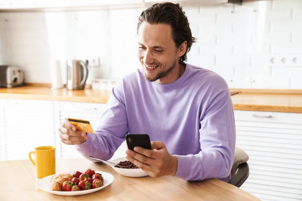 Attractive happy young man sitting at the kitchen table, having breakfast, eating cereal, shopping online with mobile phone and credit card