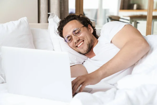 ベッドの枕の上に寝そべってる若者の笑顔ノートパソコンを使って — ストック写真