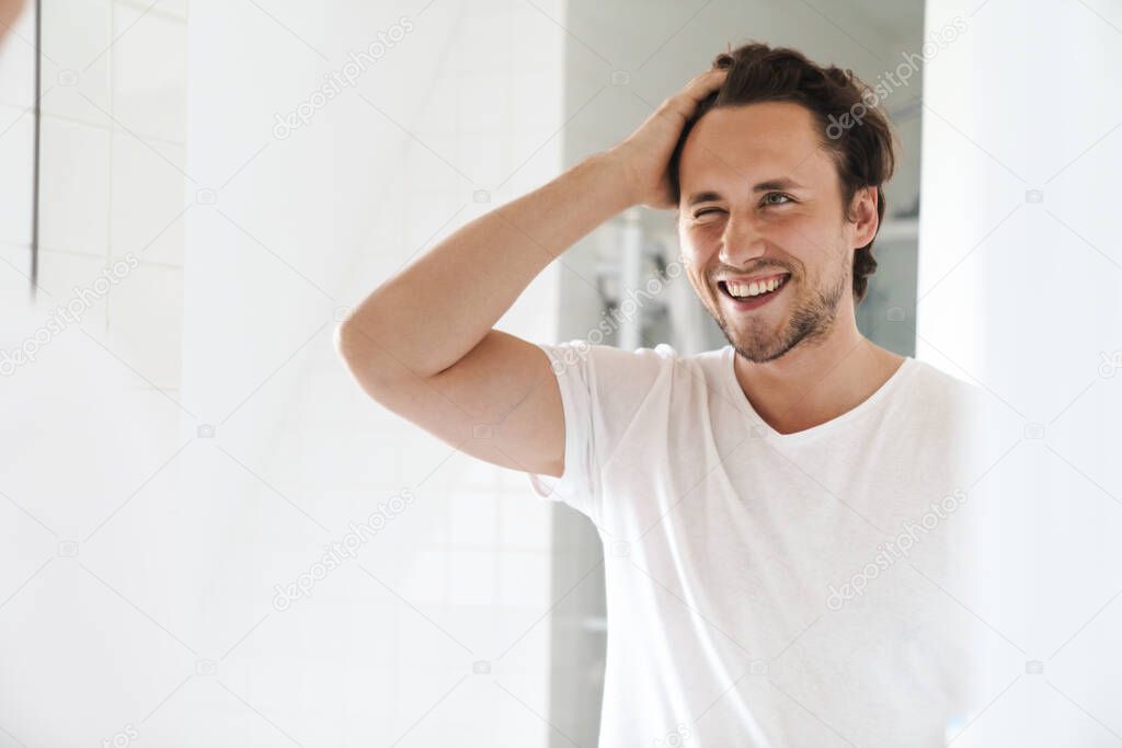 Attractive happy confident young man standing in front of the bathroom mirror, looking at himself