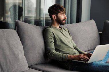 Oturma odasındaki kanepede otururken dizüstü bilgisayar kullanan rahat giyimli genç bir adam görüntüsü