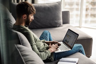 Günlük giysiler içindeki odaklanmış sakallı bir adamın oturma odasındaki kanepede otururken günlük ve dizüstü bilgisayarla çalışması.