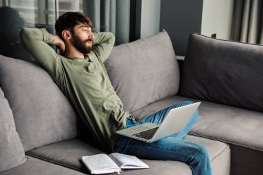 Oturma odasındaki kanepede dizüstü bilgisayarla çalışırken rahat sakallı bir adamın dinlenme görüntüsü.