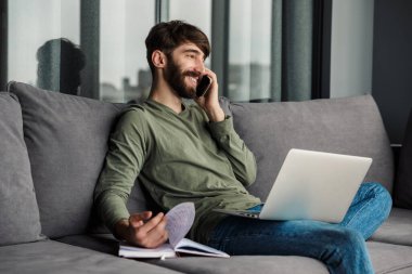 Akıllı telefondan konuşan ve oturma odasında otururken dizüstü bilgisayar kullanan sakallı bir adamın görüntüsü.