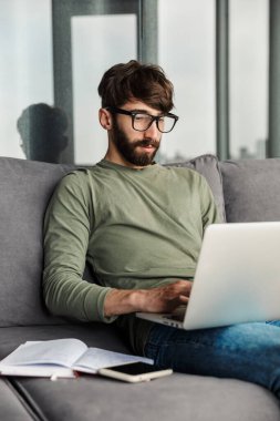 Gözlüklü, odaklanmış sakallı bir adamın oturma odasında kanepede otururken dizüstü bilgisayarla çalışması.