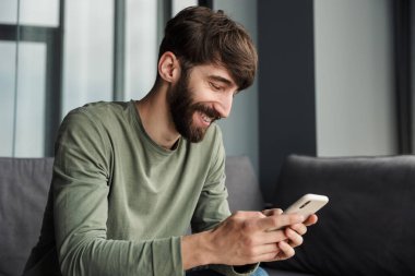 Oturma odasındaki kanepede otururken gülümseyen ve cep telefonu kullanan yakışıklı bir adam.