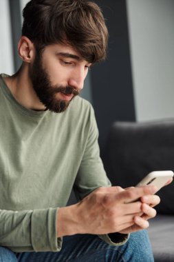 Oturma odasındaki kanepede otururken cep telefonunda daktilo çeken yakışıklı bir genç resmi.