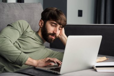 Oturma odasındaki kanepede yatarken dizüstü bilgisayar kullanan rahat giyimli genç bir adam görüntüsü