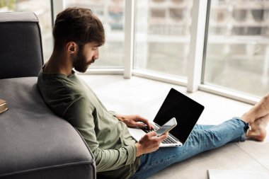 Oturma odasında yerde otururken cep telefonu ve not defteri kullanan odaklanmış genç bir adam görüntüsü