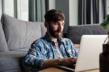 Oturma odasında yerde otururken elinde defterle çalışan sakallı bir adamın resmi.