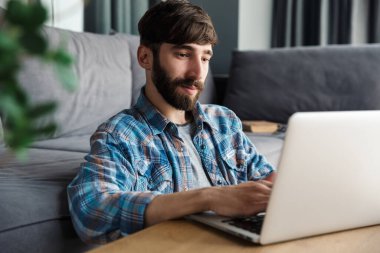 Oturma odasında yerde otururken elinde defterle çalışan sakallı adamın resmi.