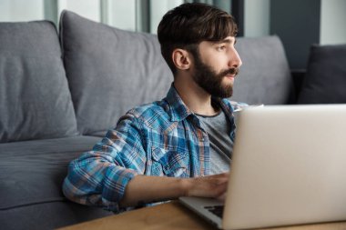 Oturma odasında yerde otururken elinde defterle çalışan sakallı bir adamın resmi.