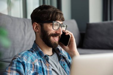 Gözlüklü, cep telefonuyla konuşan ve oturma odasında defter kullanan mutlu sakallı bir adam resmi.