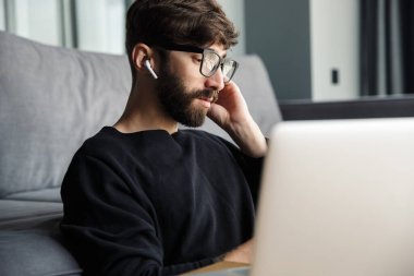 Oturma odasında otururken gözlüklü ciddi bir adamın kablosuz kulaklık ve defter kullanması.