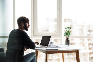 Genç ciddi bir adamın kablosuz kulaklık kullanması ve oturma odasında otururken dizüstü bilgisayarla çalışması.