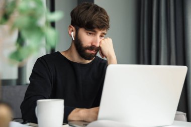 Kablosuz kulaklık kullanan ve masada otururken dizüstü bilgisayarla çalışan genç bir adam görüntüsü