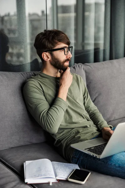 リビングルームのソファに座っている間 ノートパソコンで作業眼鏡で集中髭の男のイメージ — ストック写真