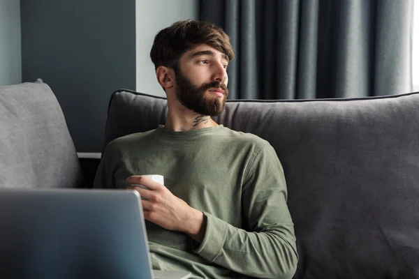 Изображение Красивого Серьезного Человека Сидящего Диване Гостиной Ноутбуком Пьющего Кофе — стоковое фото