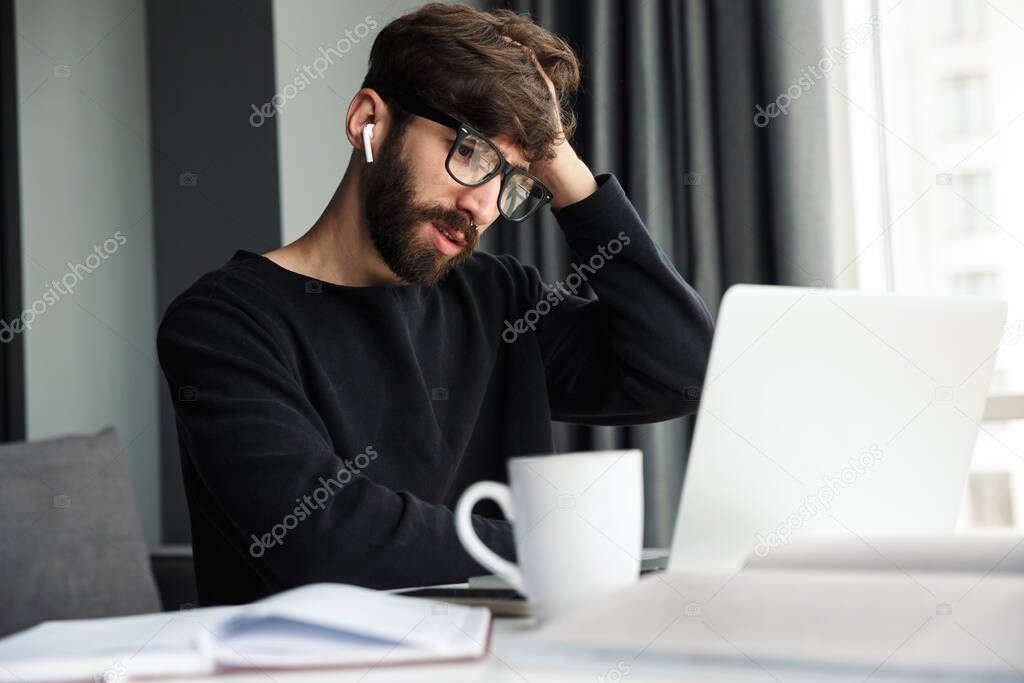 Image of young confused man using wireless earphones and working with laptop while sitting at table in living room