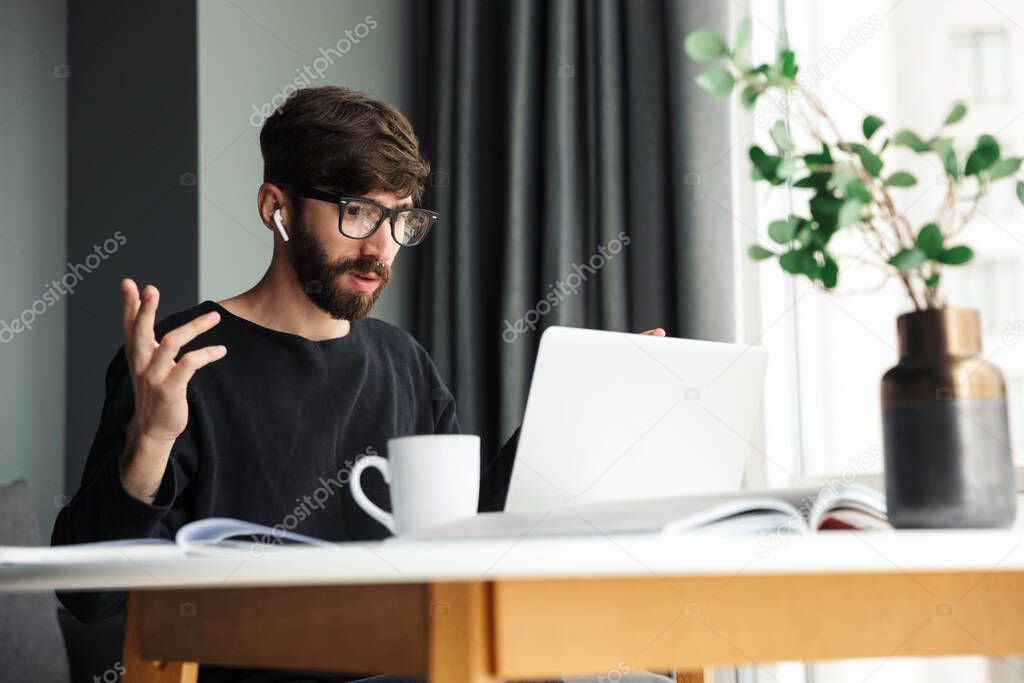 Image of young nervous man using wireless earphones and working with laptop while sitting at table in living room