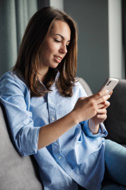Oturma odasındaki kanepede otururken gülümseyen ve cep telefonu kullanan genç bir kadın görüntüsü
