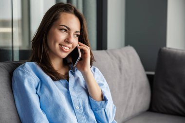 Oturma odasında otururken gülümseyen ve cep telefonuyla konuşan güzel bir kadın görüntüsü