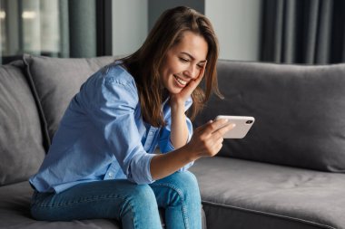 Oturma odasındaki kanepede otururken gülümseyen ve cep telefonu kullanan güzel bir kadın görüntüsü.