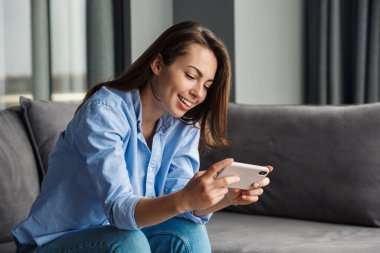 Oturma odasında otururken cep telefonuyla video oyunu oynayan güzel, neşeli bir kadın görüntüsü