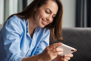 Oturma odasında otururken cep telefonuyla video oyunu oynayan güzel, neşeli bir kadın görüntüsü