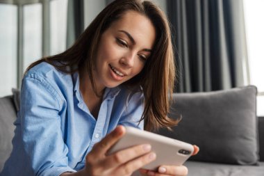 Oturma odasında otururken cep telefonuyla video oyunu oynayan güzel, neşeli bir kadın görüntüsü