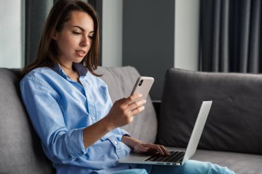 Oturma odasında kanepede otururken dizüstü bilgisayar ve cep telefonuyla çalışan genç bir kadın görüntüsü