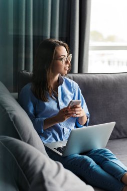 Gözlüklü mutlu bir kadının oturma odasındaki kanepede otururken cep telefonu ve dizüstü bilgisayarla çalışması.