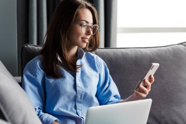 Gözlüklü mutlu bir kadının oturma odasındaki kanepede otururken cep telefonu ve dizüstü bilgisayarla çalışması.