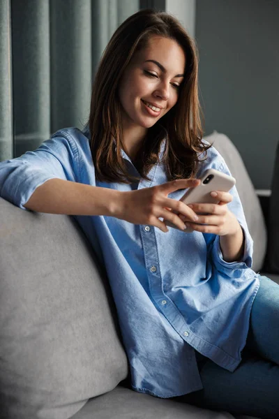 リビングルームでソファに座りながら笑顔で携帯電話を使う若い喜びの女性のイメージ — ストック写真