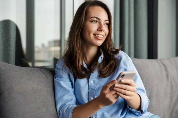 照片中年轻快乐的女人坐在客厅的沙发上笑着用手机 — 图库照片