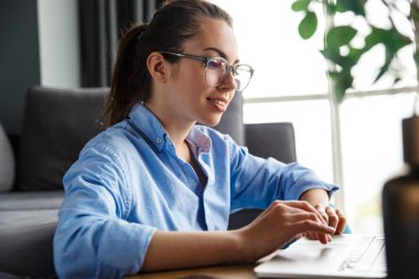 Gözlüklü, dizüstü bilgisayarla çalışan ve oturma odasında otururken gülümseyen bir kadın resmi.