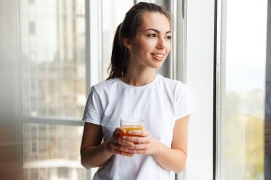 Mutlu genç bir kadın görüntüsü gülümseyen ve taze meyve suyu içen kapalı bir pencere kenarında duran.